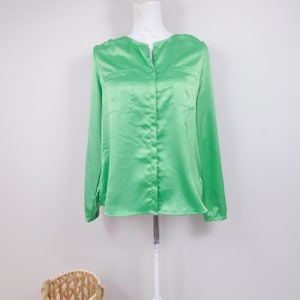 Puede incluir: Una blusa verde brillante de manga larga con dos bolsillos delanteros y cierre de botones. La blusa está hecha de un material brillante, similar al satén, y se exhibe en un maniquí blanco. La blusa tiene un diseño clásico y elegante.