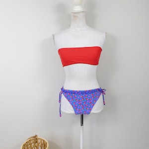 Puede incluir: Un top de bikini bandeau rojo y una parte inferior de bikini azul con estampado floral. La parte inferior del bikini tiene lazos en los lados.