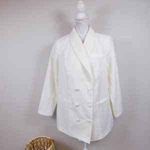 Puede incluir: Una blazer color crema de doble botonadura con seis botones a juego y dos bolsillos delanteros. La blazer se exhibe en un maniquí blanco. Una cesta tejida se encuentra en primer plano.