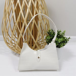 Puede incluir: Bolso blanco de piel sintética con estampado de cocodrilo y cierre dorado. El bolso tiene una sola correa y una forma ligeramente curvada.