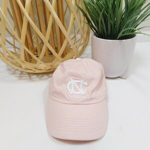 Pode incluir: Um boné de beisebol rosa com um logotipo "NC" bordado em branco e a palavra "Carolina" embaixo.