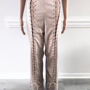 May include: Tan trousers with lace-up detailing on the sides. The high-waisted trousers have a straight leg design. The lace-up feature runs along the outer seams, adding a unique style to the garment.