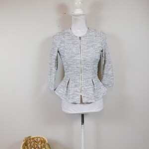 May include: A gray and white tweed jacket with a peplum waist and a zip-up front. The jacket has three-quarter sleeves and is displayed on a white mannequin. The background is a plain white wall.