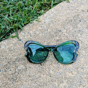 K&ouml;nnte beinhalten: Gr&uuml;ne, schmetterlingsf&ouml;rmige Sonnenbrille mit Leopardenmuster-Rahmen. Die Sonnenbrille hat gr&uuml;ne Gl&auml;ser und liegt auf einer grauen Betonfl&auml;che.