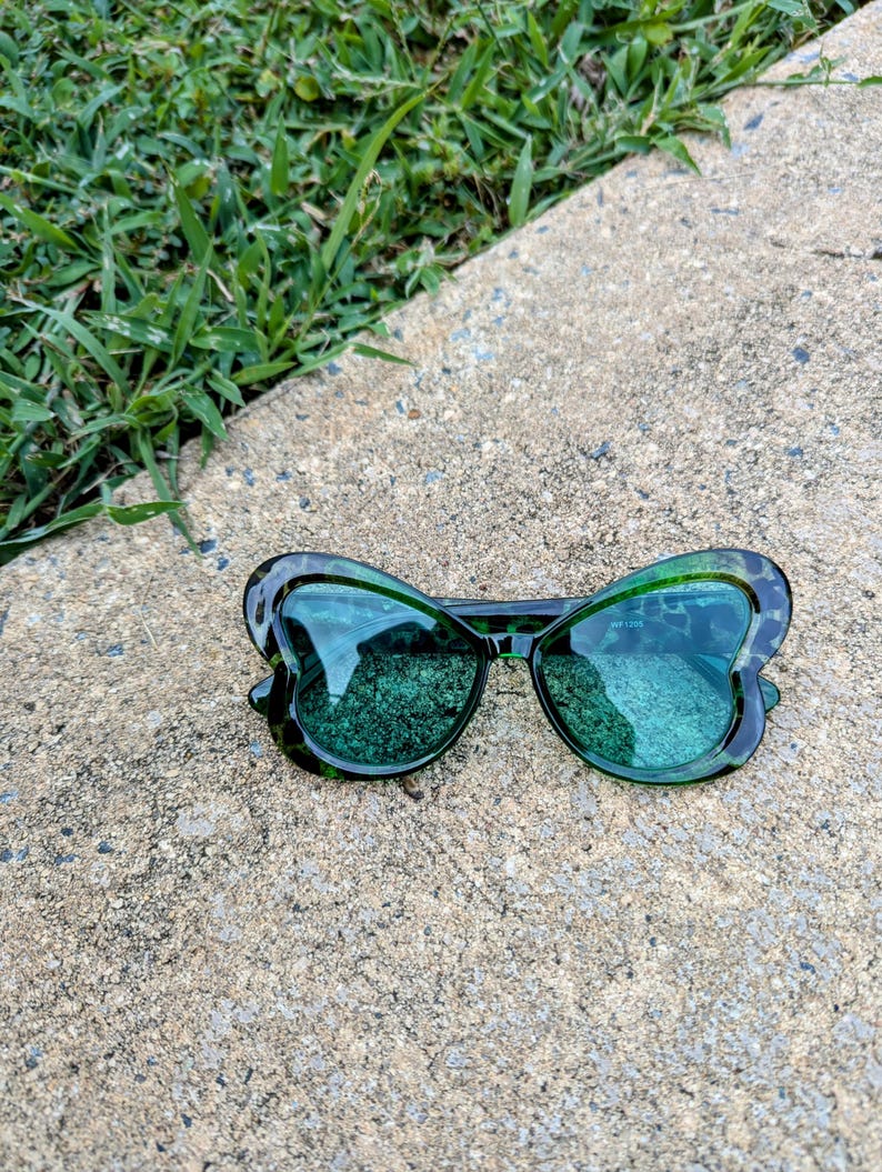 K&ouml;nnte beinhalten: Gr&uuml;ne, schmetterlingsf&ouml;rmige Sonnenbrille mit Leopardenmuster-Rahmen. Die Sonnenbrille hat gr&uuml;ne Gl&auml;ser und liegt auf einer grauen Betonfl&auml;che.