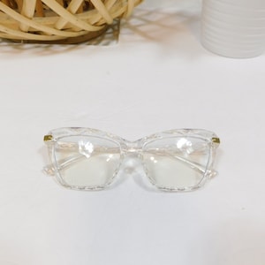 May include: Clear, square-framed spectacles with a geometric design. The spectacles have a transparent frame and lenses, with gold-coloured accents on the temples. The spectacles are set against a white background.