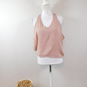 Puede incluir: Un top rosa pálido con cuello halter y escote en V se muestra en un maniquí blanco. El top tiene un diseño holgado. Una cesta tejida con asa se encuentra en la esquina inferior izquierda, añadiendo un toque rústico.