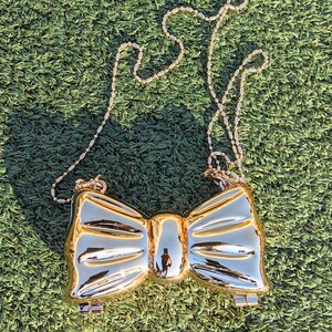 May include: A gold-colored bow-shaped clutch bag with a chain strap. The bag is lying on a green artificial grass surface.