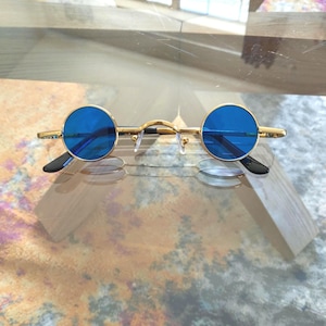 May include: Gold-framed round sunglasses with blue lenses. The glasses have a delicate gold bridge and temples, with black earpieces. The lenses are a vibrant blue color. The sunglasses are displayed on a reflective surface.