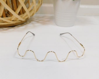 Peut inclure: Lunettes en métal doré avec une ligne de sourcil ondulée unique. Les lunettes ont une monture fine et un design distinctif et accrocheur. Le fond comprend un panier tressé et une plante en pot.