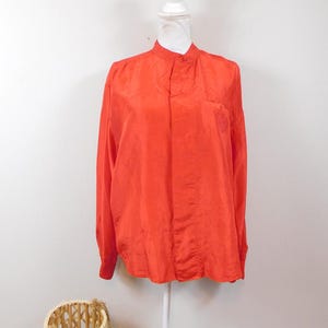 Op de afbeelding: Een lang mouwig, fel oranje overhemd met een mandarijnkraag. Het overhemd is gemaakt van een zijdeachtig materiaal en heeft een enkele knoopsluiting aan de hals.