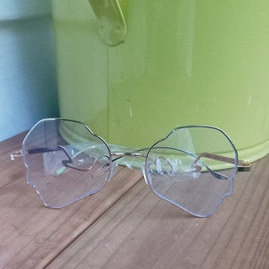 Könnte beinhalten: Eine goldene Brille mit klaren blauen Gläsern. Die Gläser sind in Form eines Gesichts mit einem abgerundeten Kinn und einer leicht spitzen Nase geformt.