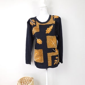 May include: A navy blue sweater with a round neckline. The sweater features a design of squares and leaves in shades of gold and brown. The sweater is displayed on a white mannequin.