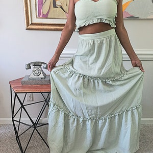 May include: A light green two-piece set featuring a strapless top and a tiered maxi skirt. The top has a ruffled edge, and the skirt has multiple ruffled tiers. The set is made of a lightweight fabric, perfect for warm weather.