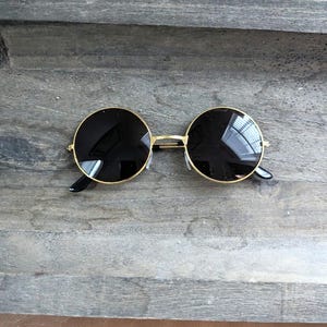 Könnte beinhalten: Runde Sonnenbrille mit goldfarbenem Rahmen und schwarzen Gläsern. Die Sonnenbrille liegt auf einer grauen Holzoberfläche.