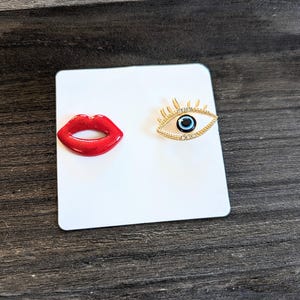 May include: A pair of earrings, one with a red lip design and the other with a blue evil eye design with rhinestones.