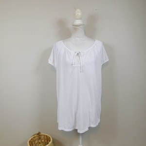 May include: A white short-sleeved top with a scoop neckline and a decorative lace insert at the chest. The top is displayed on a white mannequin. A woven basket is in the foreground.