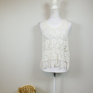 May include: Cream-coloured lace tank top with a round neckline and fringe detail at the hem. The top is displayed on a white mannequin. A woven basket is in the foreground.