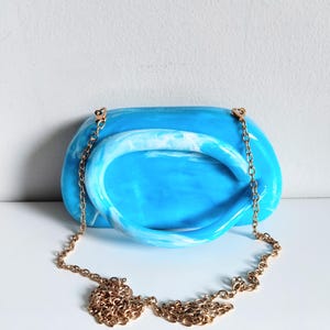 May include: A blue and white marbled resin handbag with a gold chain strap. The handbag has a rounded shape with a matching handle. The gold chain is draped in front of the bag.