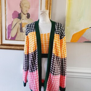May include: A multi-colored cardigan with a houndstooth pattern in black, white, pink, yellow, and green. The cardigan is open and has a long sleeve.
