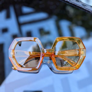 Könnte beinhalten: Orangefarbene sechseckige Sonnenbrille mit klaren Gläsern. Die Sonnenbrille ist aus Kunststoff und hat ein modernes Design.
