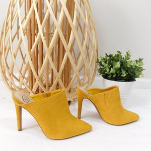 Puede incluir: Un par de mules de tacón de ante amarillo mostaza con puntera puntiaguda. Los tacones miden aproximadamente 7,5 cm de alto.