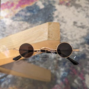 May include: Gold-toned round sunglasses with black lenses. The sunglasses have a simple, classic design.