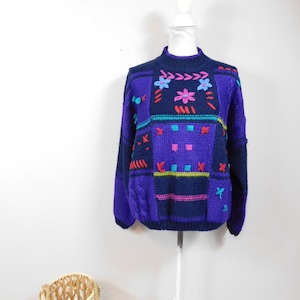 Puede incluir: Un suéter vintage azul marino y morado con coloridos patrones florales y geométricos bordados. El suéter tiene cuello alto y mangas largas. El diseño incluye acentos rosas, turquesas y amarillos, creando una estética retro vibrante.