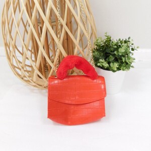 May include: A small orange faux leather purse with a crocodile pattern. The purse has a red fluffy handle and a gold chain strap.