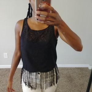 May include: A black tank top with a lace detail at the neckline and fringe trim at the hemline. The top is paired with white pants. The image is taken in front of a mirror.