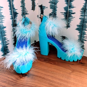 May include: A pair of bright turquoise platform sandals featuring a chunky block heel, a lug sole, and a deep blue insole. The footwear is adorned with fluffy, light blue and white feathers across the toe strap and the ankle strap.