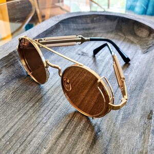 May include: Gold-framed sunglasses with brown lenses. The sunglasses have a unique design with spring-like details on the temples and a bridge. The lenses are round and the frame is a shiny gold color. The sunglasses are resting on a gray wooden surface.