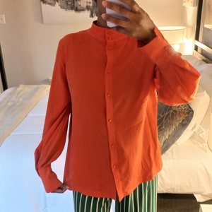 Könnte beinhalten: Ein orangefarbenes langärmliges Hemd mit Stehkragen und Knopfleiste. Dazu eine Hose mit grün-weißen Streifen im weiten Bein-Stil. Die Aufnahme zeigt die gesamte Person.