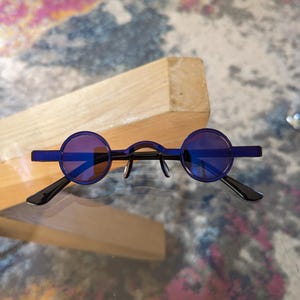 May include: A pair of round, dark blue sunglasses with black frames. The sunglasses are resting on a wooden surface.