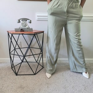 May include: Light green high-waisted pants with a gathered waistband and wide legs. The pants are paired with white pointed-toe shoes. A black geometric side table with a wooden top and a vintage telephone are also visible.