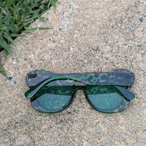K&ouml;nnte beinhalten: Gr&uuml;ne Sonnenbrille mit einem Schildpattmuster am Rahmen. Die Gl&auml;ser sind dunkelgr&uuml;n.