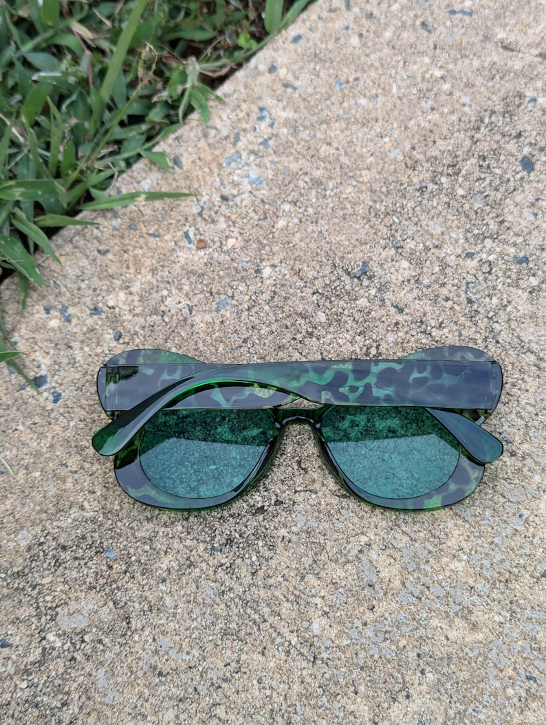 K&ouml;nnte beinhalten: Gr&uuml;ne Sonnenbrille mit einem Schildpattmuster am Rahmen. Die Gl&auml;ser sind dunkelgr&uuml;n.