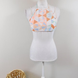 May include: A halter top with a geometric pattern in shades of orange, blue, and white. The top is displayed on a white mannequin. A woven basket is in the lower left corner.