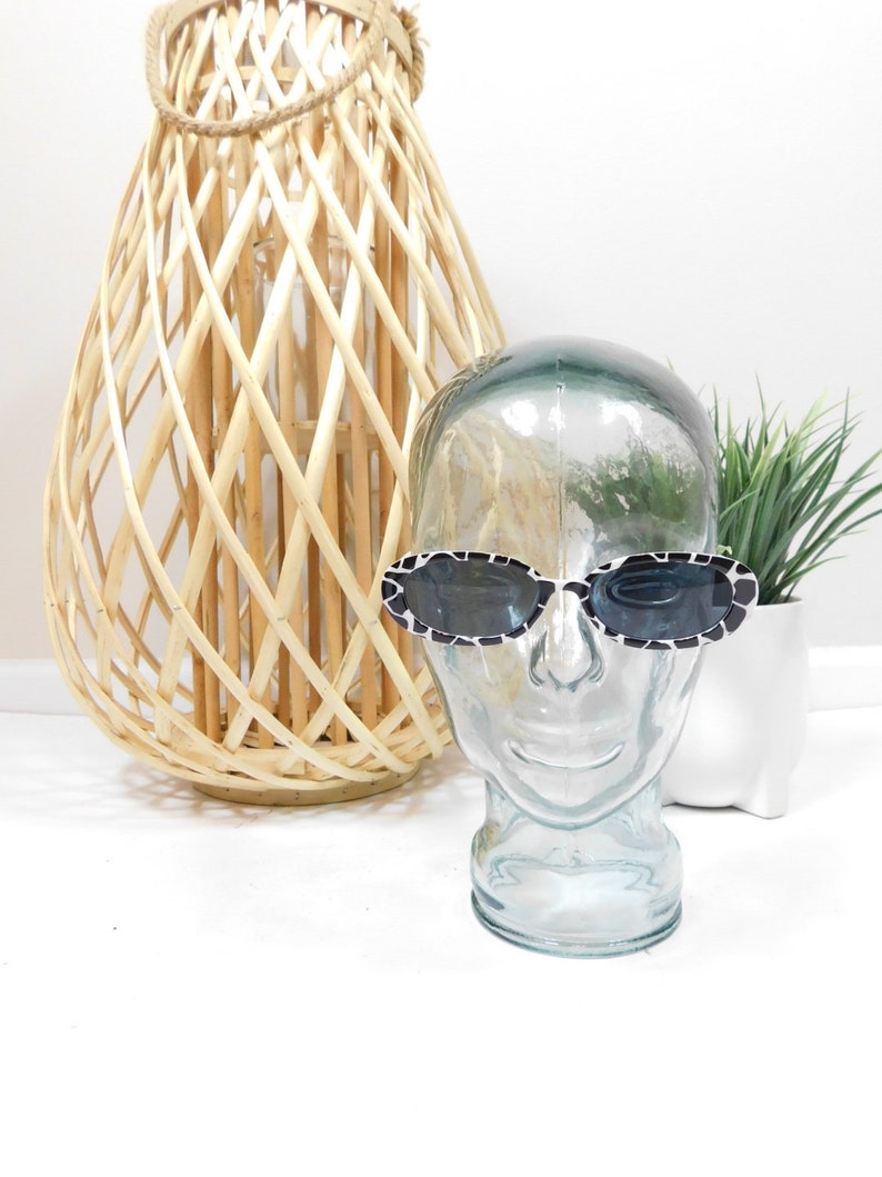 May include: Black and white patterned sunglasses with a clear plastic frame on a glass mannequin head. The sunglasses are on a white surface.
