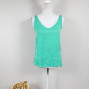 May include: A sleeveless, turquoise-colored tank top with a V-neck is displayed on a white mannequin. The top has a slightly textured appearance. A wicker basket is visible in the lower left corner.