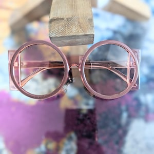 May include: A pair of round, pink-tinted sunglasses with a geometric frame design. The sunglasses are made of plastic and have a modern, stylish look.