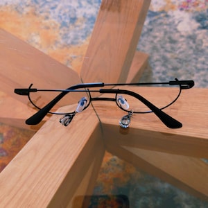 May include: Black-rimmed eyeglasses with a delicate, minimalist design. The glasses feature a half-rim style with a thin frame and teardrop-shaped charms dangling from the lenses. The glasses are set on a wooden surface.