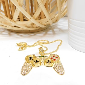 May include: A gold-coloured pendant necklace with a video game controller design. The controller is encrusted with clear and red rhinestones, with black accents on the buttons. The necklace chain is also gold-coloured.