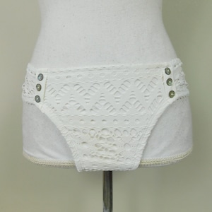 Puede incluir: Braga de bikini blanca de crochet con botones decorativos. La braga tiene un borde festoneado y un patrón de encaje.