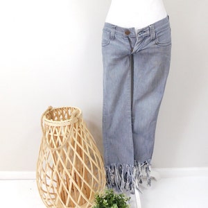 May include: Light blue denim jeans with a frayed hem, displayed on a mannequin. A woven basket with a rope handle and a small potted plant are in the foreground. The jeans have a button and zipper closure.