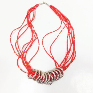 Può includere: Una collana multifilo realizzata con perline di vetro rosse e un ciondolo in metallo argentato. Il ciondolo è composto da più anelli collegati.