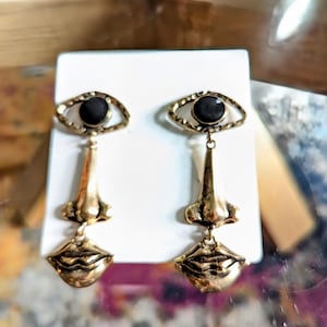 May include: A pair of gold-toned earrings featuring a face design. Each earring has an eye with a black stone, a nose, and a pair of lips. The earrings are displayed on a white card.