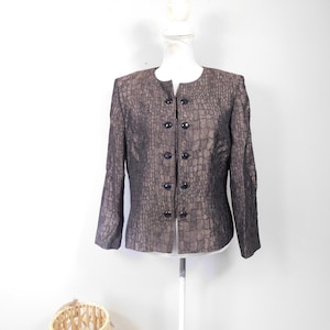 Könnte beinhalten: Eine braun-schwarz strukturierte Jacke mit rundem Ausschnitt und schwarzen Knöpfen. Die Jacke wird auf einer weißen Schaufensterpuppe präsentiert. Der Stoff hat ein reptilienartiges Muster.