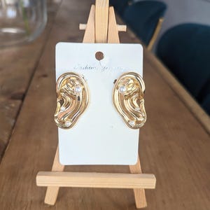May include: Gold-tone earrings shaped like an ear with a pearl accent. The earrings are displayed on a white card with the text "Fashion Jewelry".