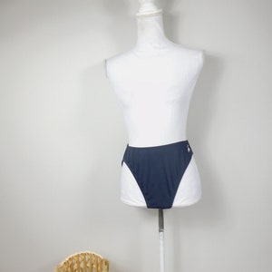 Puede incluir: Braguita de bikini azul marino de cintura alta sobre un maniquí blanco. La braguita tiene un diseño sencillo con un pequeño logotipo en el lateral. Una cesta de mimbre está en primer plano.